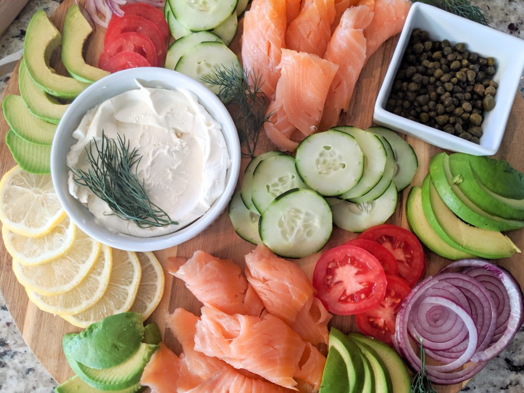 Bagel and lox toppings. Including: cream cheese smear, capers, red onion, of course lox, cucumber, tomato, avocado, lemon, and dill. All of these toppings nicely placed on a charcuterie cutting board.